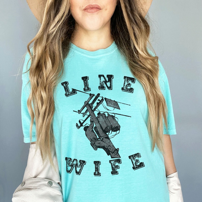 May include: A light blue t-shirt with a graphic of a lineman working on a power line. The text "LINE WIFE" is printed in black letters on the shirt.
