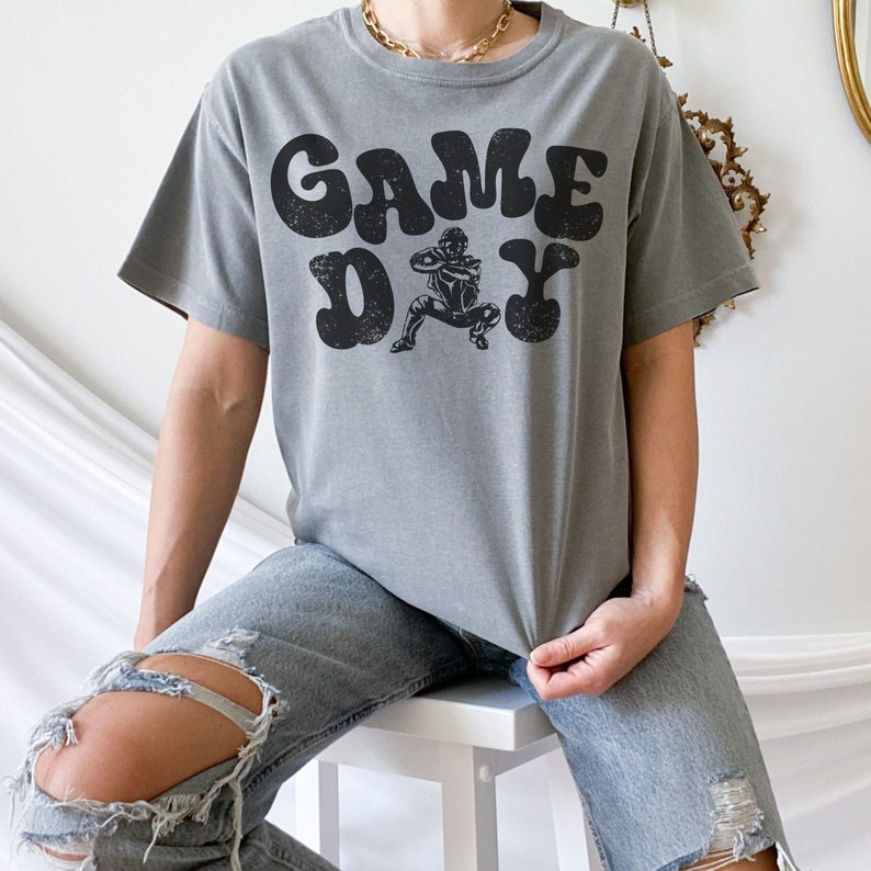 May include: A grey t-shirt with a vintage-style graphic of a football player and the text "GAME DAY".