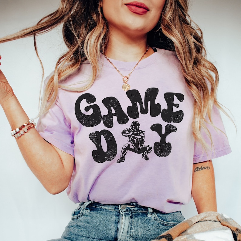 May include: A lavender t-shirt with a black graphic that says "GAME DAY" in a retro font. A cartoon football player is in the middle of the text.