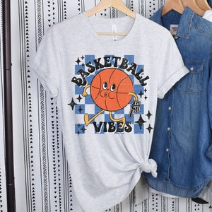 May include: A gray t-shirt with a retro-style basketball graphic and the text "Basketball Vibes". The basketball is orange and has a cartoon face. The text is in blue and white.