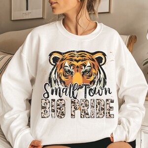 Pu&ograve; includere: Felpa bianca con una grafica di un viso di tigre rovinato e il testo "Small Town Big Pride" in nero su uno sfondo con stampa leopardata.