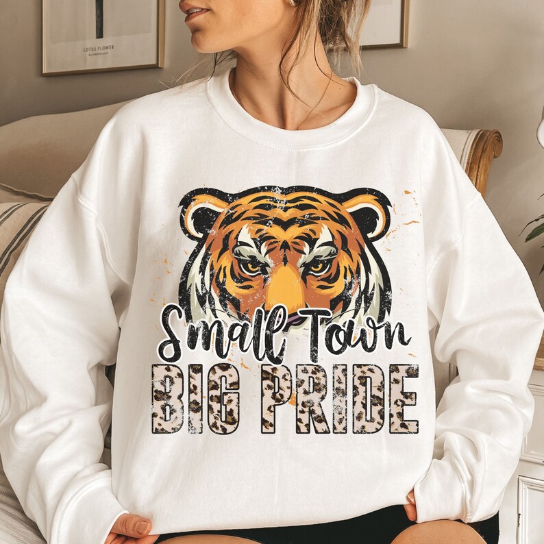 Pu&ograve; includere: Felpa bianca con una grafica di un viso di tigre rovinato e il testo "Small Town Big Pride" in nero su uno sfondo con stampa leopardata.