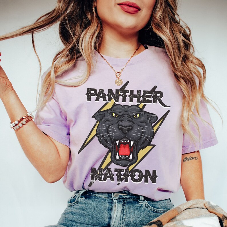 Op de afbeelding: Een paars T-shirt met een zwarte panter afbeelding en de tekst "PANTHER NATION" in zwart en geel. De panter brult met zijn bek open en heeft gele ogen.