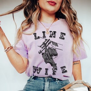 May include: A purple t-shirt with the text "LINE WIFE" in black with a distressed look. The shirt features a graphic of a lineman working on a power line.