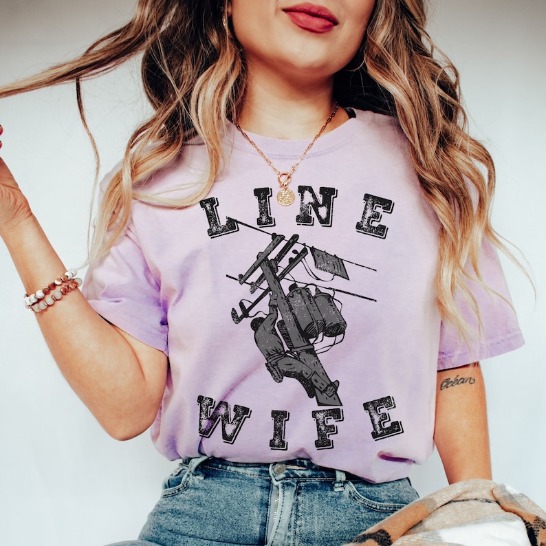 May include: A purple t-shirt with the text "LINE WIFE" in black with a distressed look. The shirt features a graphic of a lineman working on a power line.