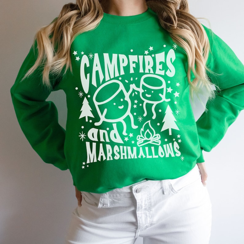 Puede incluir: Sudadera verde con un dise&ntilde;o gr&aacute;fico blanco de dos malvaviscos de dibujos animados, una fogata y el texto "Campfires and Marshmallows".