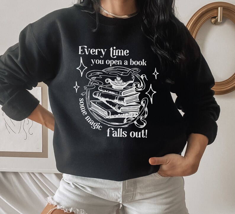 May include: Black sweatshirt with white graphic text: "Every time you open a book & some magic falls out!" The design features a stack of books, a genie lamp, and stars. A casual, long-sleeved top for book lovers.