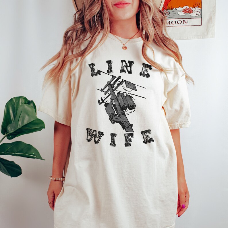 May include: A white t-shirt with the text "LINE WIFE" in black lettering. The shirt features a black and white illustration of a person working on a power line.