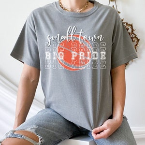 Puede incluir: Camiseta gris con las palabras "small town big pride" en letras blancas y un dise&ntilde;o de baloncesto naranja y blanco. La camiseta es de estilo casual.