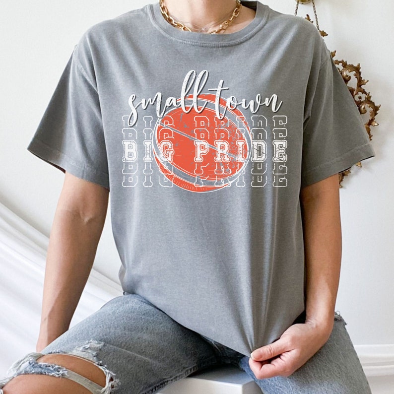 Puede incluir: Camiseta gris con las palabras "small town big pride" en letras blancas y un dise&ntilde;o de baloncesto naranja y blanco. La camiseta es de estilo casual.