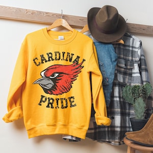 Op de afbeelding: Geel sweatshirt met een rode kardinaalvogel afbeelding en de tekst "CARDINAL PRIDE" in zwart.
