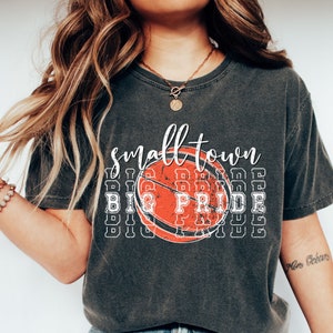 Puede incluir: Camiseta gris oscuro con las palabras "small town" en escritura blanca sobre un gr&aacute;fico de baloncesto naranja desgastado. Debajo del baloncesto, las palabras "BIG PRIDE" est&aacute;n apiladas en un patr&oacute;n repetitivo.