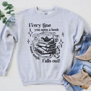May include: A light gray sweatshirt with the text "Every time you open a book some magic falls out!" in black. The graphic features a stack of books, a genie lamp, and stars.