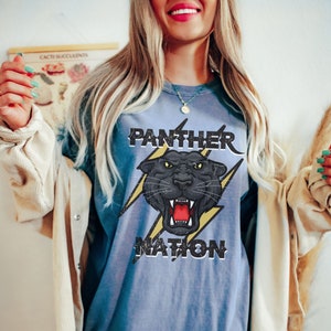 Op de afbeelding: Een blauw T-shirt met een zwarte panter afbeelding en de tekst "Panther Nation" in een verweerd lettertype.