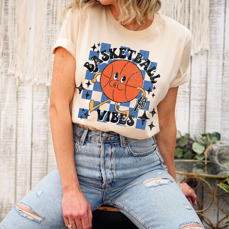 May include: A cream colored t-shirt with a cartoon basketball graphic. The basketball has a face and is holding a basketball. The text "Basketball Vibes" is printed on the shirt in a retro font.