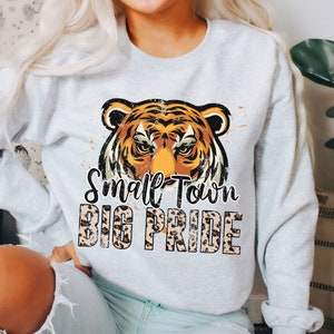 Pu&ograve; includere: Una felpa grigia con una grafica di un viso di tigre e il testo "Small Town Big Pride" in un font a stampa leopardata.