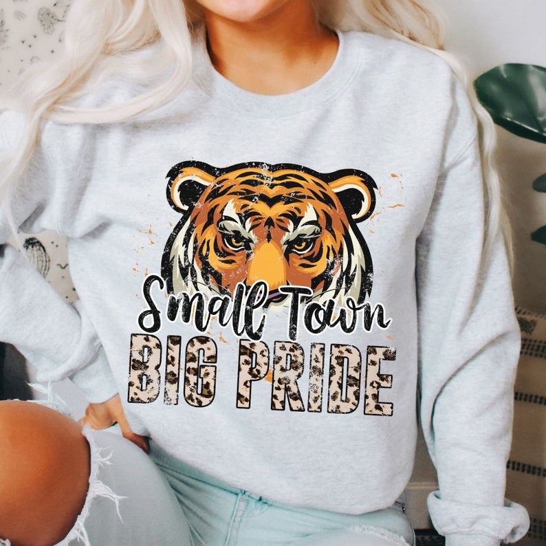 Pu&ograve; includere: Una felpa grigia con una grafica di un viso di tigre e il testo "Small Town Big Pride" in un font a stampa leopardata.