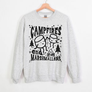 Puede incluir: Sudadera gris con un dise&ntilde;o gr&aacute;fico negro de dos malvaviscos de dibujos animados tom&aacute;ndose de la mano frente a una fogata con el texto "Campfires and Marshmallows".