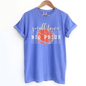 Puede incluir: Una camiseta azul b&iacute;garo con las mangas enrolladas. La camiseta presenta un gr&aacute;fico de baloncesto con las palabras "small town BIG PRIDE" en letras blancas y naranjas. La camiseta est&aacute; colgada de una percha de madera.