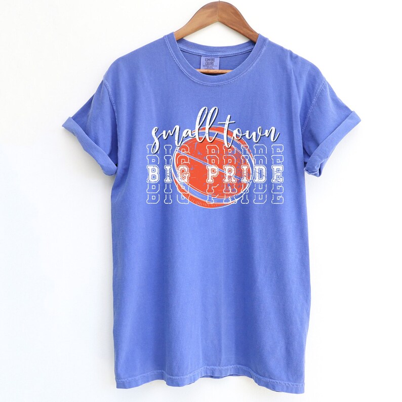 Puede incluir: Una camiseta azul b&iacute;garo con las mangas enrolladas. La camiseta presenta un gr&aacute;fico de baloncesto con las palabras "small town BIG PRIDE" en letras blancas y naranjas. La camiseta est&aacute; colgada de una percha de madera.