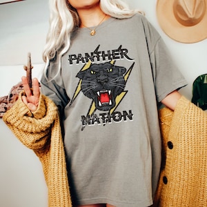 Op de afbeelding: Een grijs oversized t-shirt met een zwarte panter afbeelding en de tekst "PANTHER NATION" in een verweerd lettertype.
