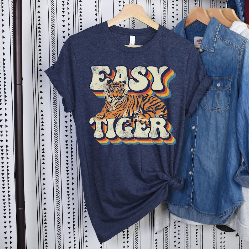 May include: A navy blue t-shirt featuring a retro-style graphic of a tiger and the text "EASY TIGER" in rainbow colours.