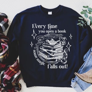 May include: Navy blue sweatshirt with white text and graphic. The text reads "Every time you open a book some magic falls out!" The graphic features a stack of books, a genie lamp, and stars. A cozy, casual style.