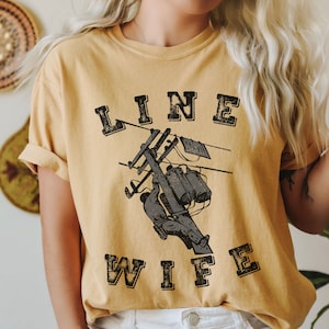 May include: A yellow t-shirt with the text "LINE WIFE" in black with a distressed look. The shirt features a graphic of a lineman working on a power line.