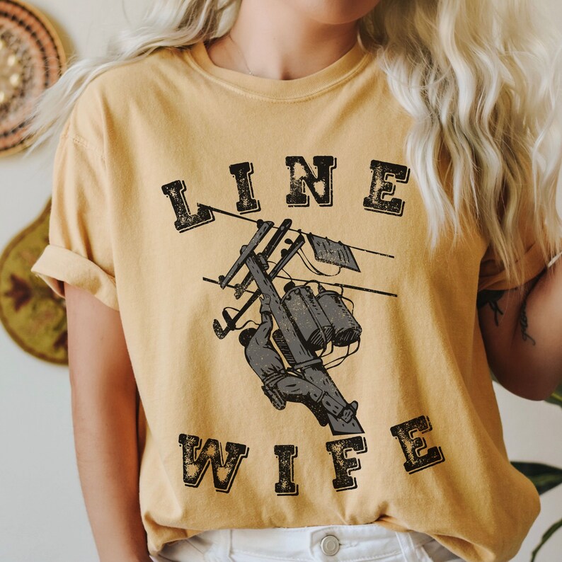 May include: A yellow t-shirt with the text "LINE WIFE" in black with a distressed look. The shirt features a graphic of a lineman working on a power line.