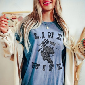 May include: A blue t-shirt with a distressed graphic of a lineman working on a power line and the text "LINE WIFE".