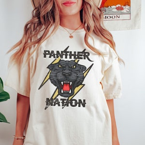 Op de afbeelding: Een wit T-shirt met een zwarte panter afbeelding en de tekst "Panther Nation" in een vintage stijl.