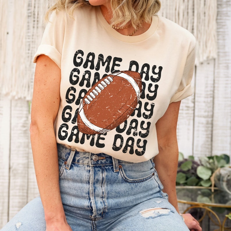 May include: A beige t-shirt with the text "GAME DAY" repeated in a retro font. A brown and white football graphic is centered on the shirt.