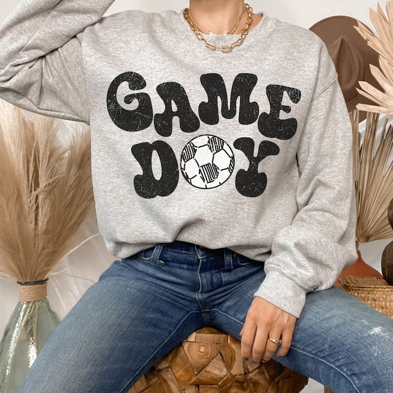 May include: A gray sweatshirt with the text "GAME DAY" in a retro font. A black and white soccer ball is centered between the words.