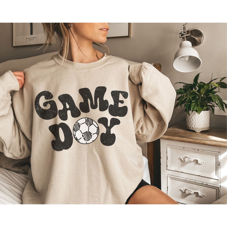 May include: A beige sweatshirt with a black graphic print that says "GAME DAY" with a soccer ball in the middle of the word "DAY".