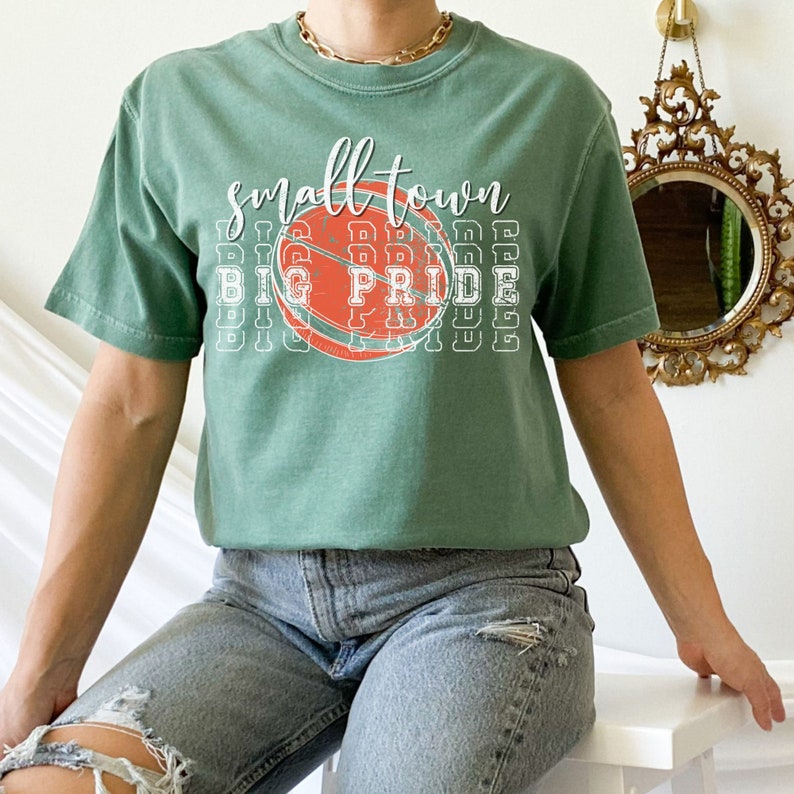Puede incluir: Una camiseta verde de manga corta con las palabras "small town" sobre un gr&aacute;fico de baloncesto. Debajo, las palabras "big pride" se repiten verticalmente. La camiseta se combina con unos vaqueros azules.
