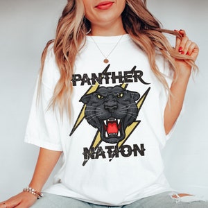 Op de afbeelding: Wit T-shirt met een zwarte panter afbeelding en de tekst "PANTHER NATION" in zwart en geel.