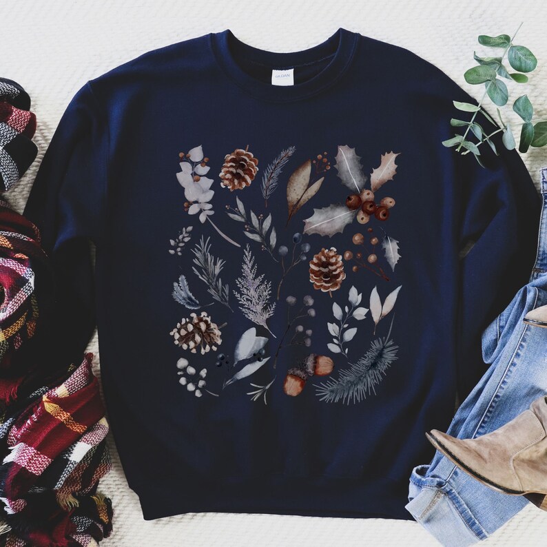 Pu&ograve; includere: Una felpa blu navy con un design ad acquerello di fogliame invernale, tra cui pigne, bacche e foglie.