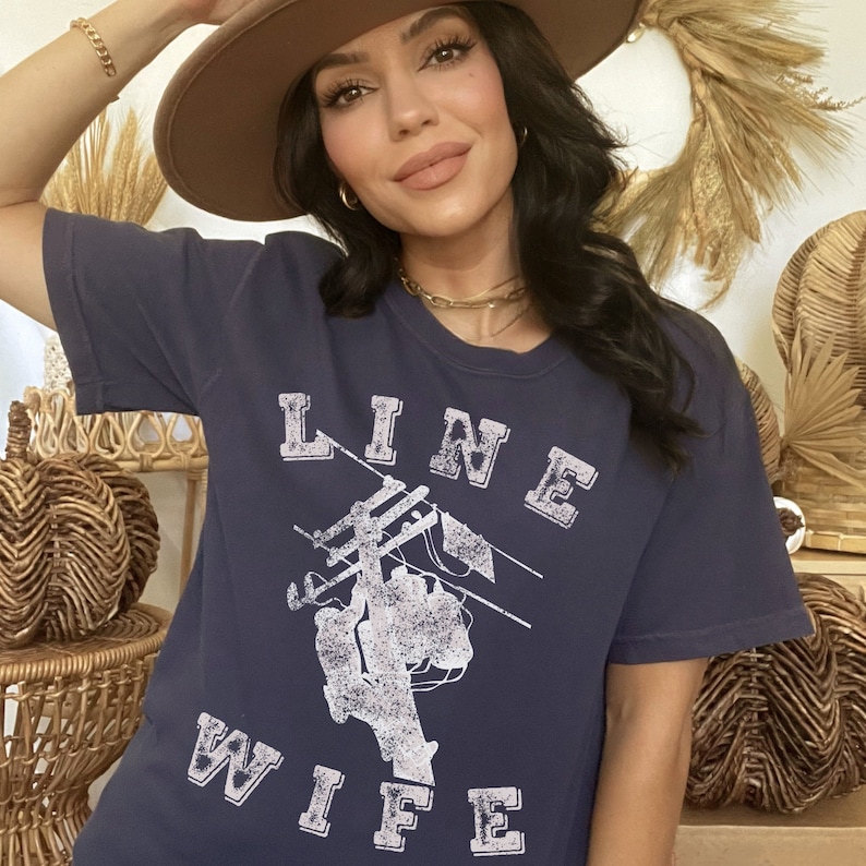 May include: A navy blue t-shirt with a white graphic of a person working on a power line. The text "LINE WIFE" is printed in white on the shirt.
