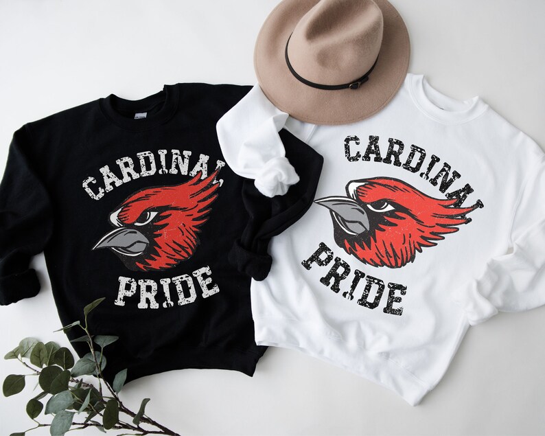 Op de afbeelding: Twee sweatshirts met een rode kardinaalvogel afbeelding en de tekst "Cardinal Pride" in een verweerd lettertype. Een sweatshirt is zwart en de andere is wit.