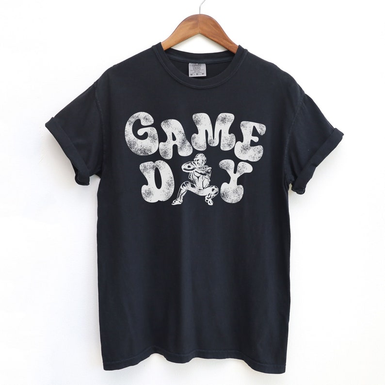 May include: Black t-shirt with a white graphic print of a football player in a vintage style font that says "GAME DAY".