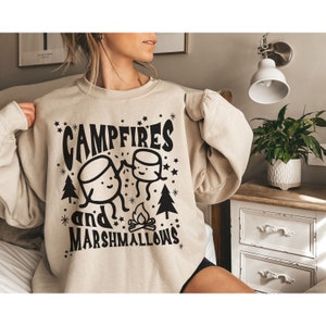Puede incluir: Una sudadera beige con un dise&ntilde;o gr&aacute;fico negro que dice "Campfires and Marshmallows" con dos malvaviscos de dibujos animados y una fogata.