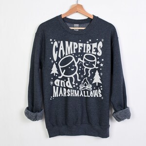 Puede incluir: Una sudadera gris oscuro con un dise&ntilde;o gr&aacute;fico blanco que dice "Campfires and Marshmallows" con una imagen de dibujos animados de dos malvaviscos y una fogata.