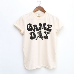 May include: A cream coloured t-shirt with a black graphic of a football player and the text "GAME DAY" in a retro font.