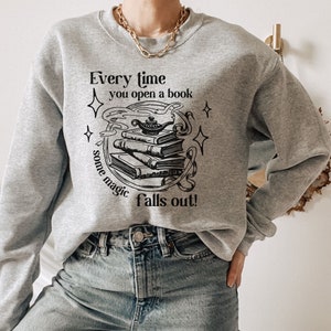 May include: A light gray sweatshirt with the text "Every time you open a book some magic falls out!" in black. The graphic features a stack of books, a genie lamp, and stars. The sweatshirt is a crew neck style.