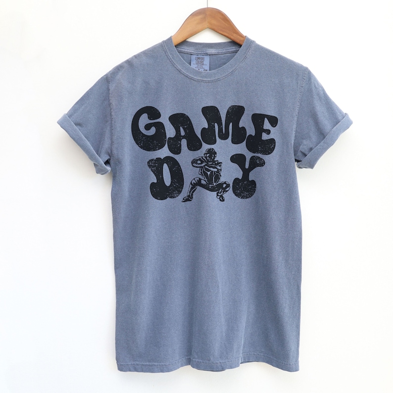 May include: A blue t-shirt with a black graphic print of a football player in a retro style font that says "GAME DAY".
