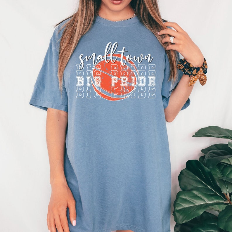 Puede incluir: Camiseta azul con la frase "small town big pride" en blanco y naranja, con un gr&aacute;fico de baloncesto naranja. La camiseta tiene un estilo casual y es c&oacute;moda.