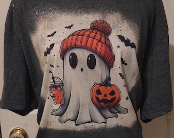 May include: Dark gray t-shirt featuring a cartoon ghost wearing an orange knit cap and holding a pumpkin and a drink. The ghost is surrounded by bats, creating a Halloween theme.