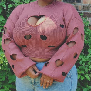 Puede incluir: Un suéter de color rosa polvoriento con recortes en forma de corazón en las mangas y el pecho. El suéter tiene una textura acanalada y un cuello redondo. Las mangas son largas y acampanadas. La persona lleva vaqueros azules.