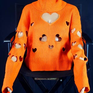 May include: A bright orange ribbed knit sweater featuring a high turtleneck collar and multiple heart-shaped cutouts distributed across the front and down both long sleeves. The garment has a straight hemline and long sleeves with ribbed cuffs.