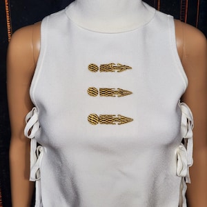 May include: A white sleeveless top with a turtleneck collar and gold embroidered details. The top features three horizontal gold designs and white tie closures on the sides. The fabric appears to be a knit material.